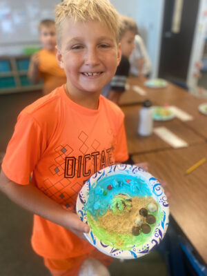 Boy in orange shirt with relief map created with icing and other edible treats on a paper plate.
