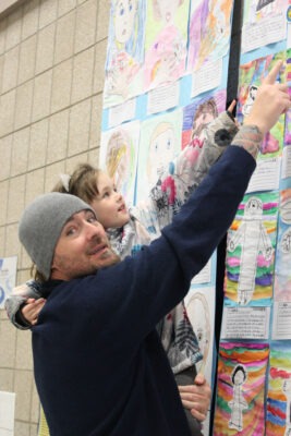 A man lifts his daughter so she can point out her artwork.